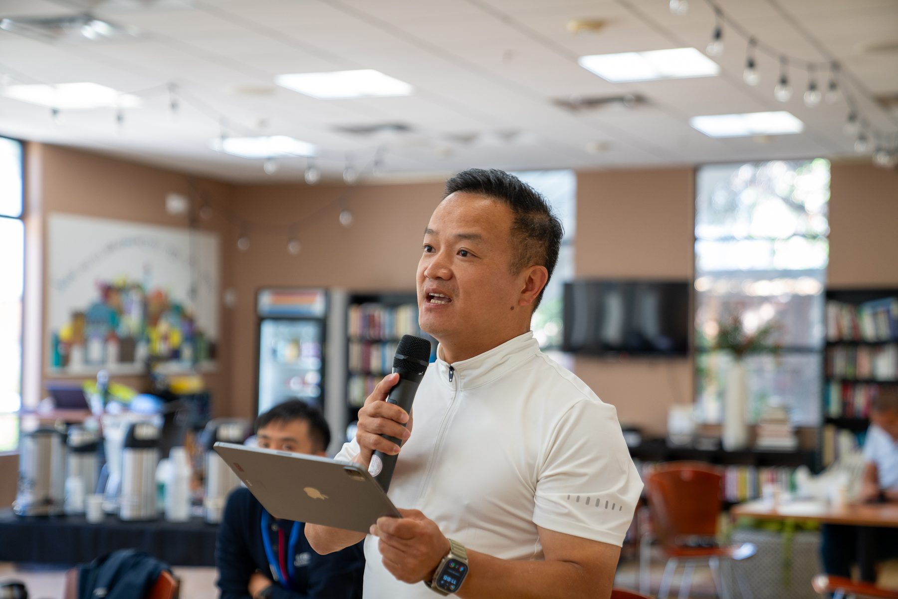 James TJ Lee speaking with a handheld microphone and tablet in a library classroom.