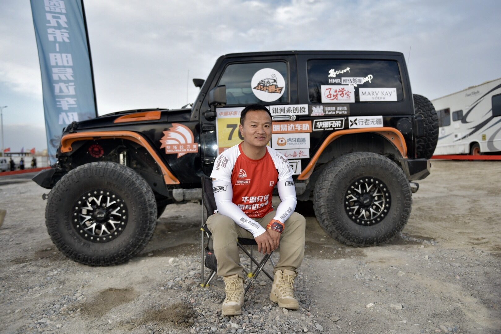 James TJ Lee seated before a black-and-orange off-road vehicle at a rally bivouac.