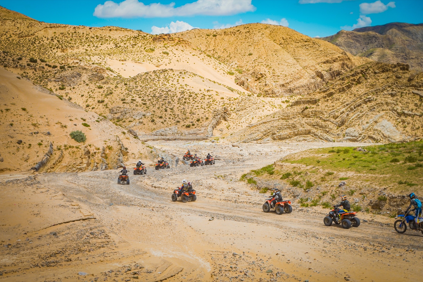 A line of helmeted Little Knights riders crossing a broad rocky valley on ATVs and a motorcycle